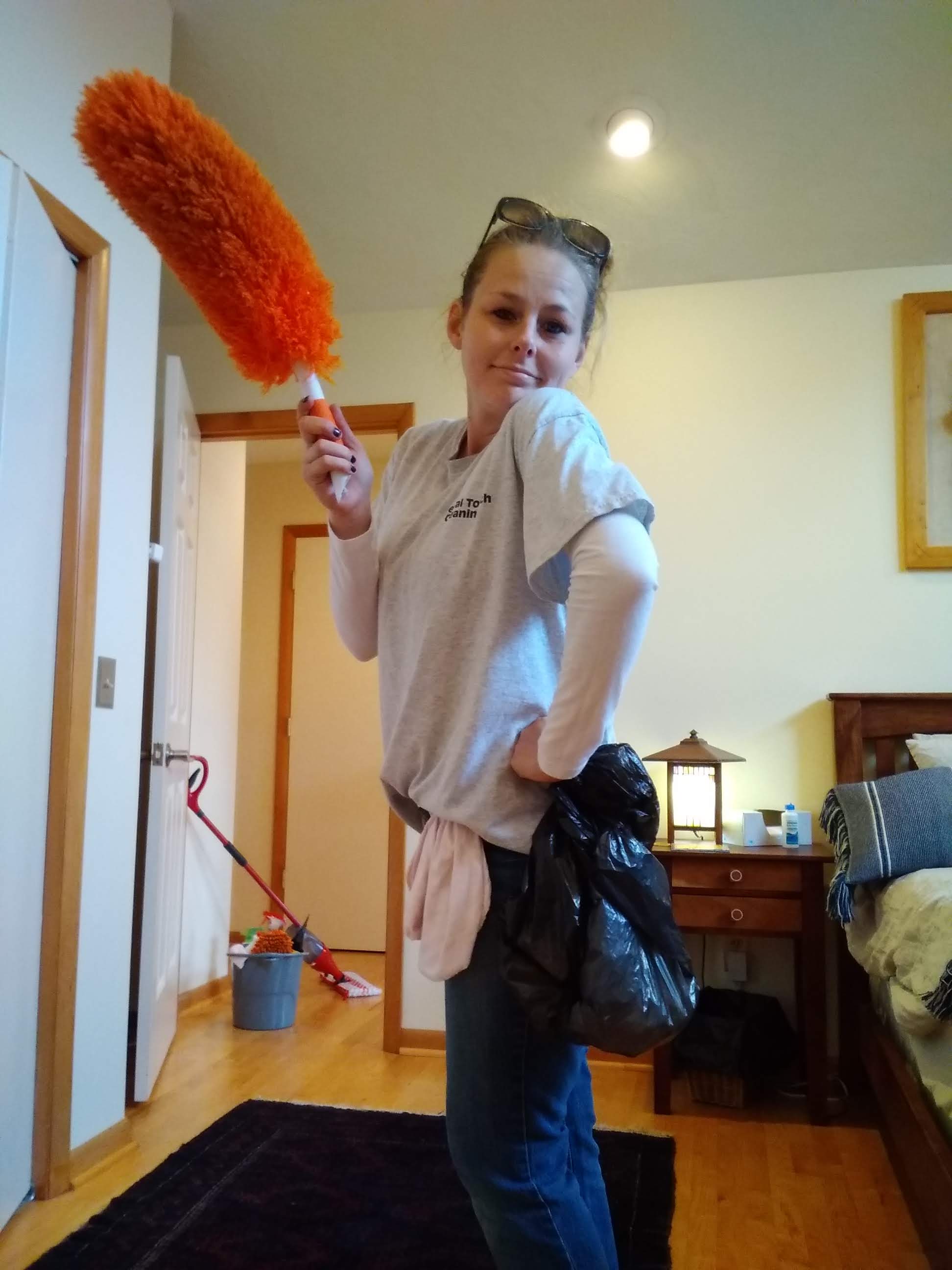 A woman in a denim outfit vacuuming a light-colored carpet in a bright living room with a yellow sofa and a potted plant.